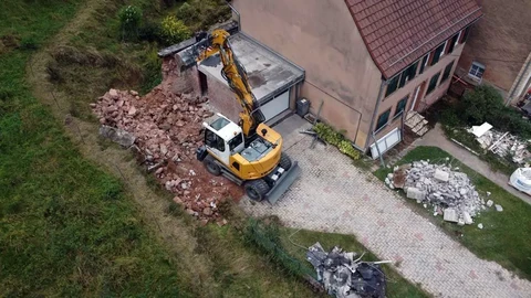 Chantier de démolition en cours avec pelleteuse hydraulique jaune sur chenilles opérant près d'une maison résidentielle à toit de tuiles rouges
